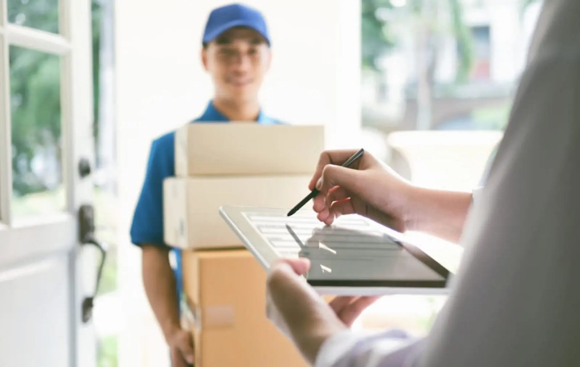 A delivery person stands at another person's doorstep carrying 4 boxes while the recipient signs for the packages on a tablet