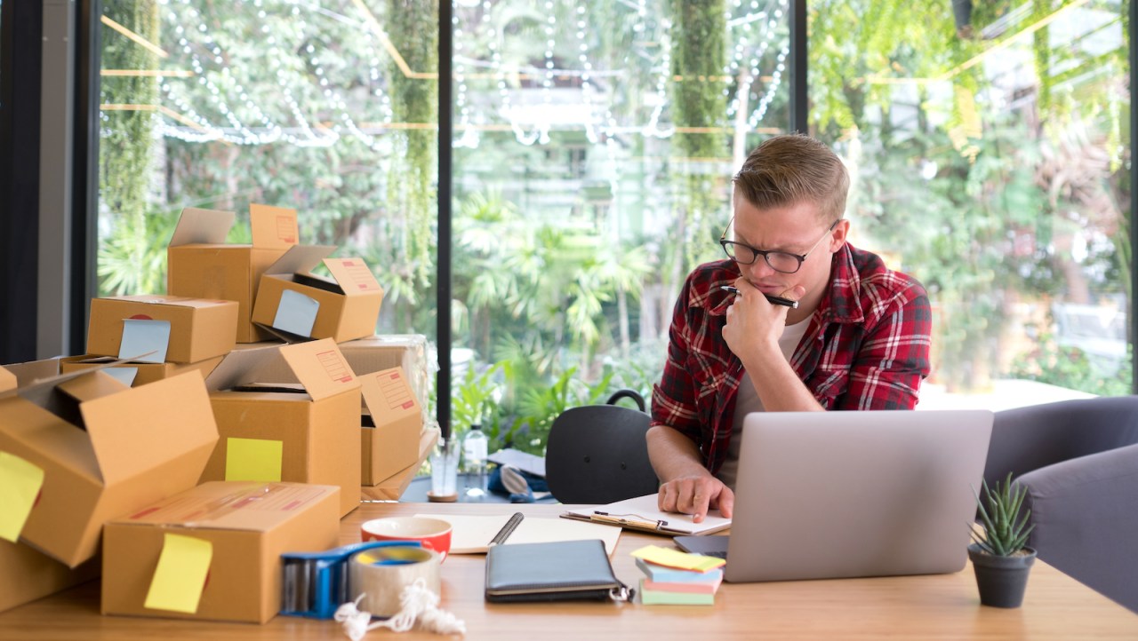 A person in a red plaid shirt sits in front of a laptop, with a pile of empty boxes on their right