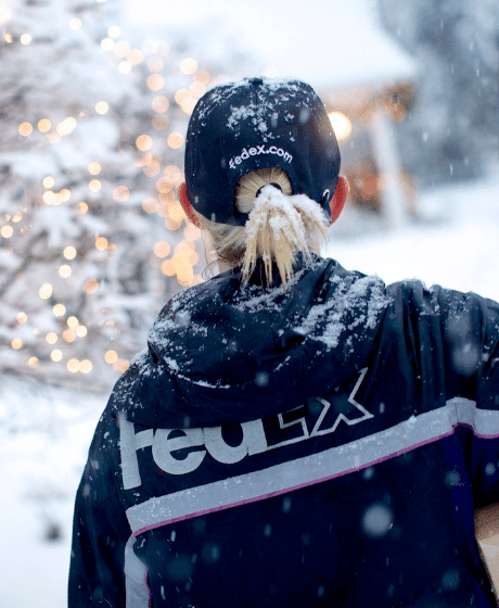A blonde woman delivers a package in the snow, wearing a black winter coat and black baseball cap that both have FedEx's logo printed on the back