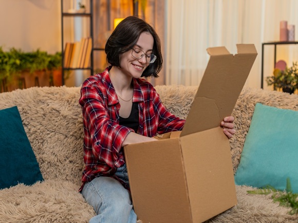 Young happy woman opens package on home couch, smiling with satisfaction while looking at new delivered items. Girl reacts with joy and excitement, enjoying shopping result and pleasant surprise _id2633797521