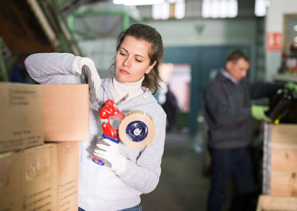 Female employee working at warehouse on artisanal olive oil factory, preparing carton boxes for delivery on market, sealing with sticky tape_id1698408826