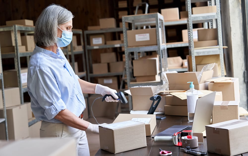 Older mature female online store small business owner worker wearing face mask packing package scanning postal drop shipping ecommerce retail order in box preparing delivery parcel in stock warehouse. _id1841388892