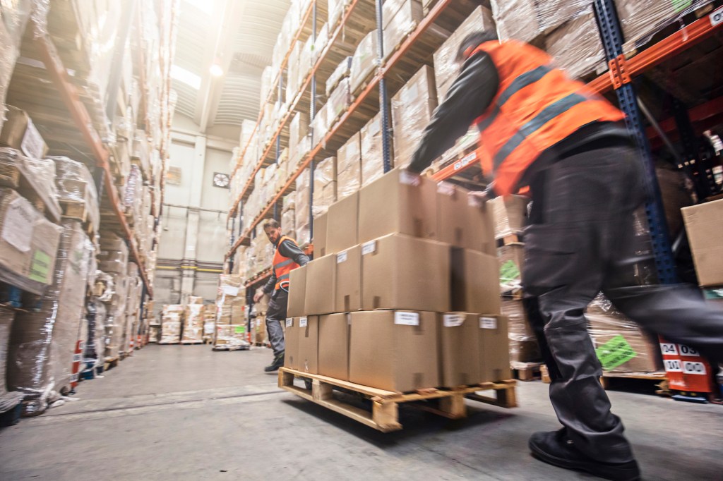 A worker in an orange construction vest pushes a pallet of boxes through a fully-stocked warehouse aisle