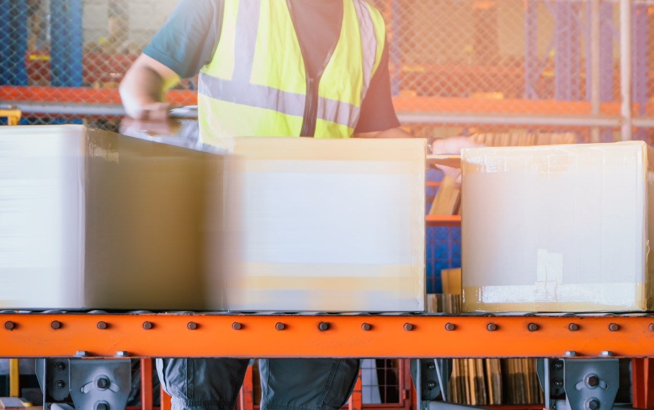 Three boxes move along a conveyor belt, helped along by a person wearing a yellow safety vest.