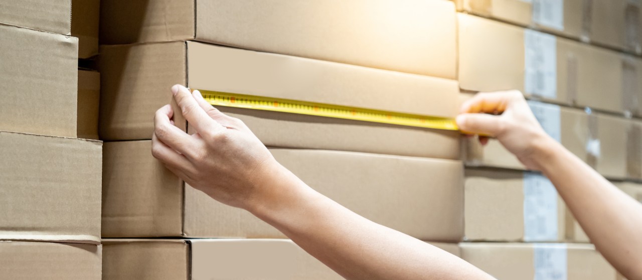 Hands measure a one of many identical long cardboard boxes with a yellow measuring tape