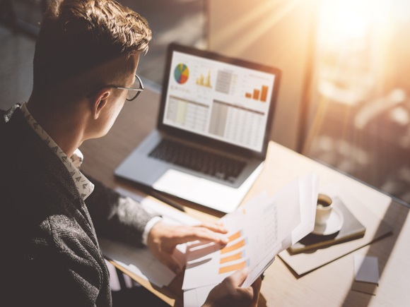 Young finance market analyst in eyeglasses working at sunny office on laptop while sitting at wooden table.Businessman analyze document in his hands.Graphs and diagramm on notebook screen.Blurred_id608938430