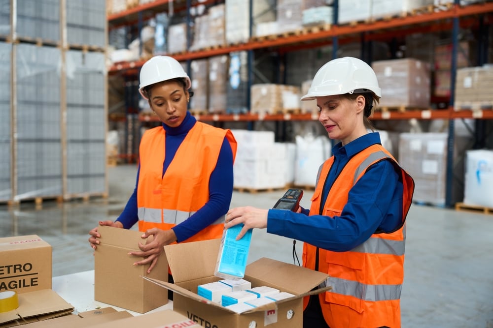 Two women workers wearing safety gear packing products into cardboard boxes and scanning them in a warehouse_id2710119145
