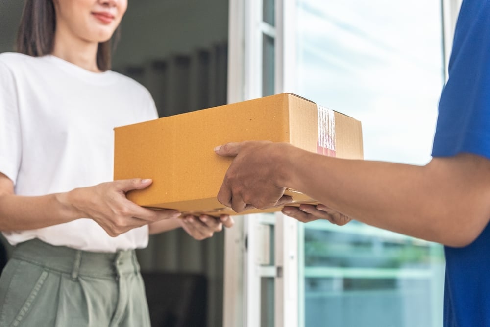 Close up delivery man hands wearing blue uniform send a cardboard box to customer in front of the customer's house with sunset flare background.