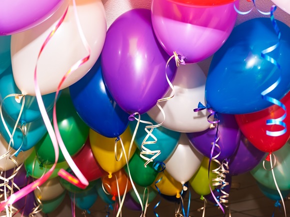 A cluster of colorful helium balloons with curling ribbons floats under a ceiling during an indoor celebration, likely a party or festive event _id2650999821