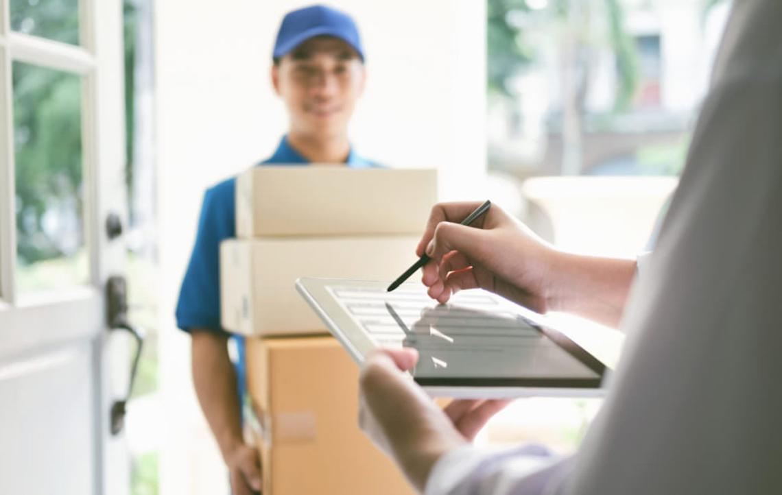 A delivery person stands at another person's doorstep carrying 4 boxes while the recipient signs for the packages on a tablet