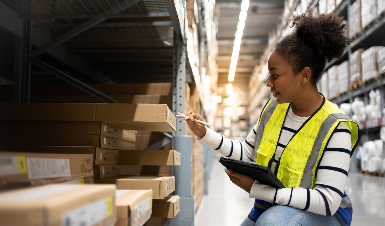 A warehouse associate in a neon yellow safety vest inspects boxes of inventory on a warehouse shelf.