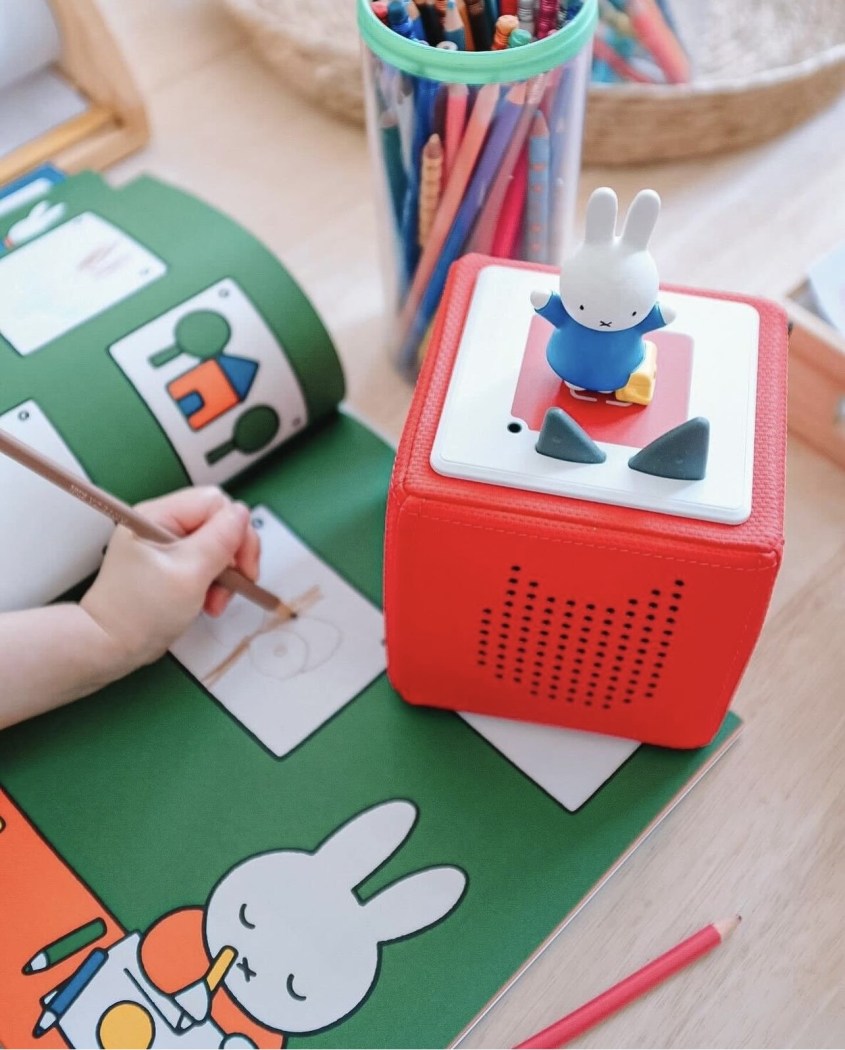A child colors in a green coloring book while listening to a red Tonies box with a white rabbit tonie on top of it