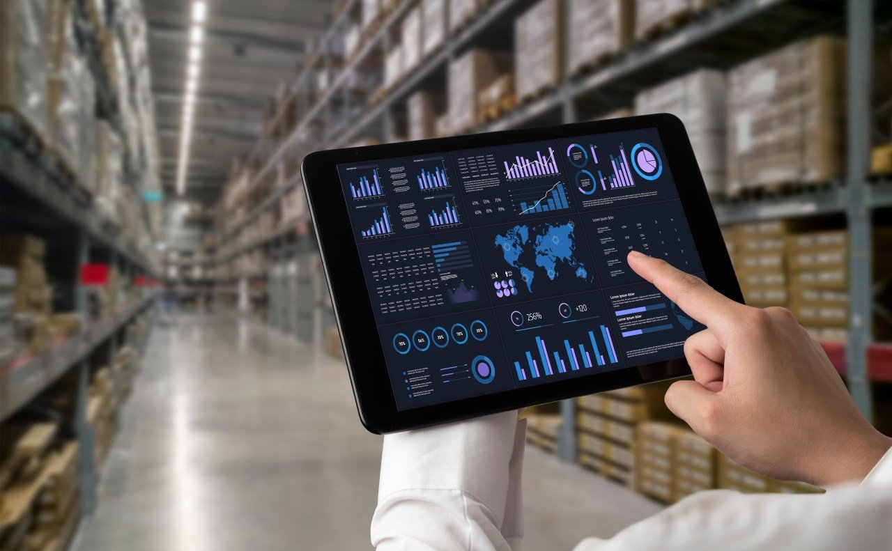 Person analyzing warehouse analytics dashboard on tablet in a large fulfillment center, showcasing inventory tracking, supply chain metrics, and data visualization tools.
