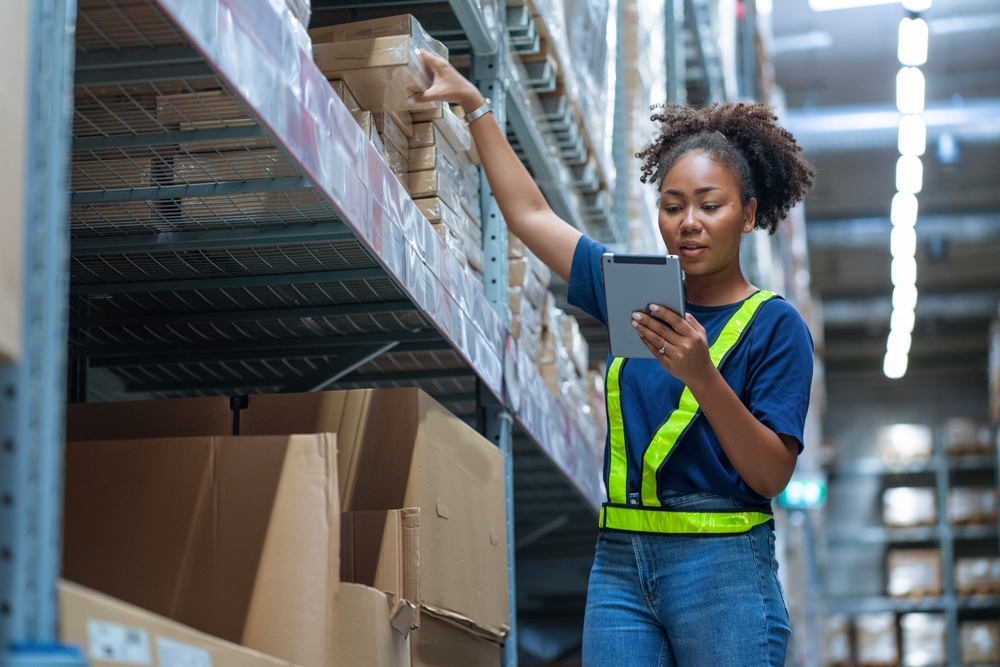 A woman holding a tablet checking inventory and orders from customers, picking up boxes from warehouse shelves